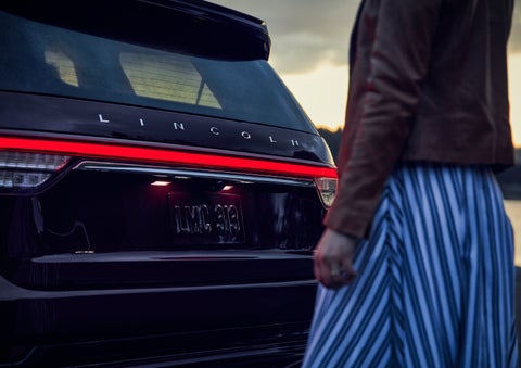 A person is shown near the rear of a 2024 Lincoln Aviator® SUV as the Lincoln Embrace illuminates the rear lights | Empire Lincoln of Huntington in Huntington NY